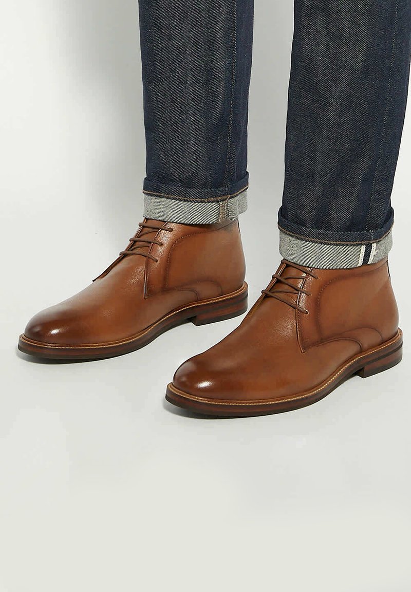 Brown leather ankle boots with a smooth finish, featuring a rounded toe, lace-up design, and a sturdy sole. Worn with dark denim jeans.