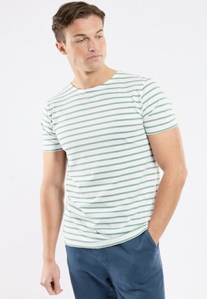 Homme portant un T-shirt blanc à rayures horizontales vertes et un pantalon bleu, regardant vers le bas et sur sa gauche, une main dans la poche.