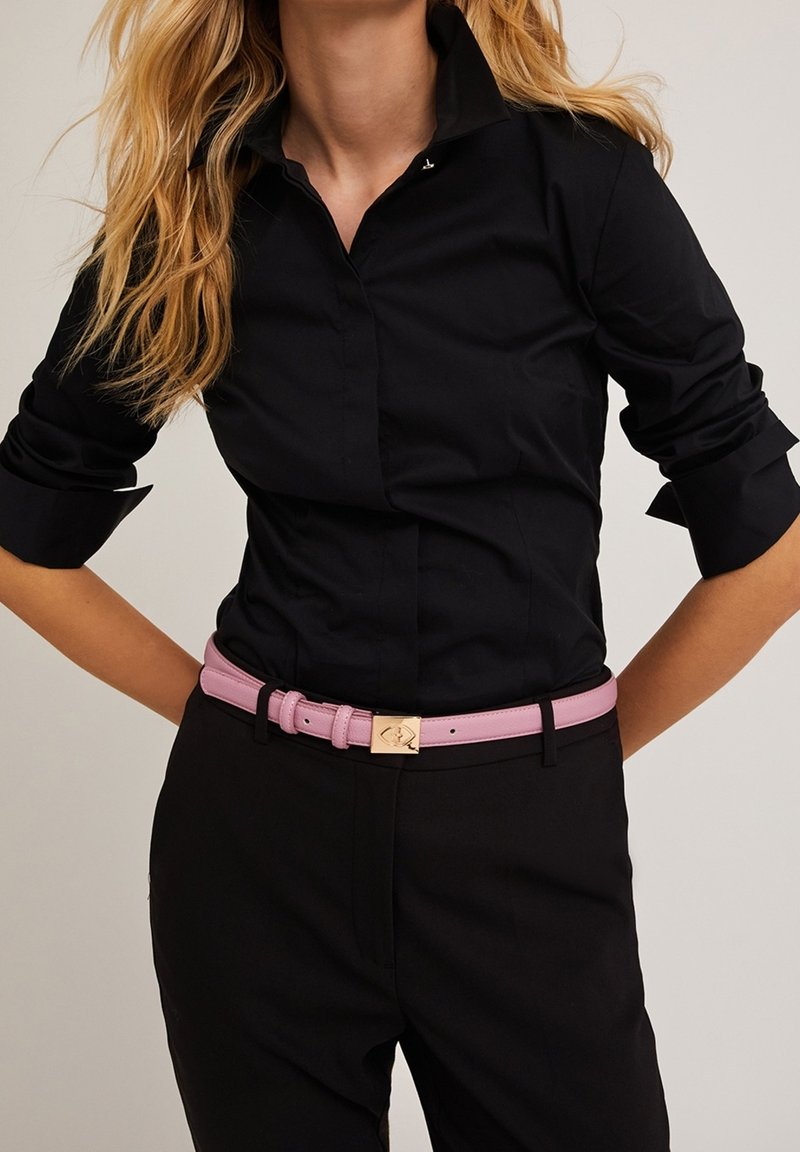 Femme portant une chemise noire cintrée à boutons avec les manches retroussées et un pantalon noir, accessoirisée d'une ceinture rose à boucle dorée.
