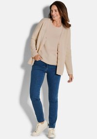 Beige strukturierte Strickjacke über einem passenden Oberteil, kombiniert mit blauen Skinny-Jeans und weißen Sneakers. Verfügt über Knöpfe und eine lässige Passform.
