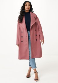 Donna con lunghi capelli ricci che indossa un cappotto doppiopetto rosa polveroso, un dolcevita blu navy, jeans azzurri arrotolati e tacchi neri con cinturino alla caviglia.