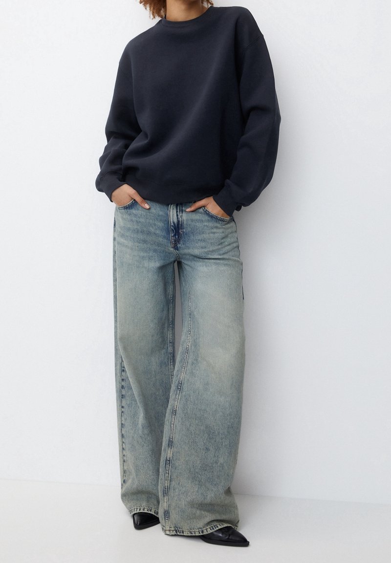 Personne portant un sweat-shirt bleu marine, un jean large délavé, des chaussures noires pointues, debout les mains dans les poches contre un mur blanc.