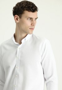 Camicia bianca a maniche lunghe con tessuto strutturato, chiusura frontale a bottoni e colletto classico, con bottoni color avorio semplici e una vestibilità su misura.