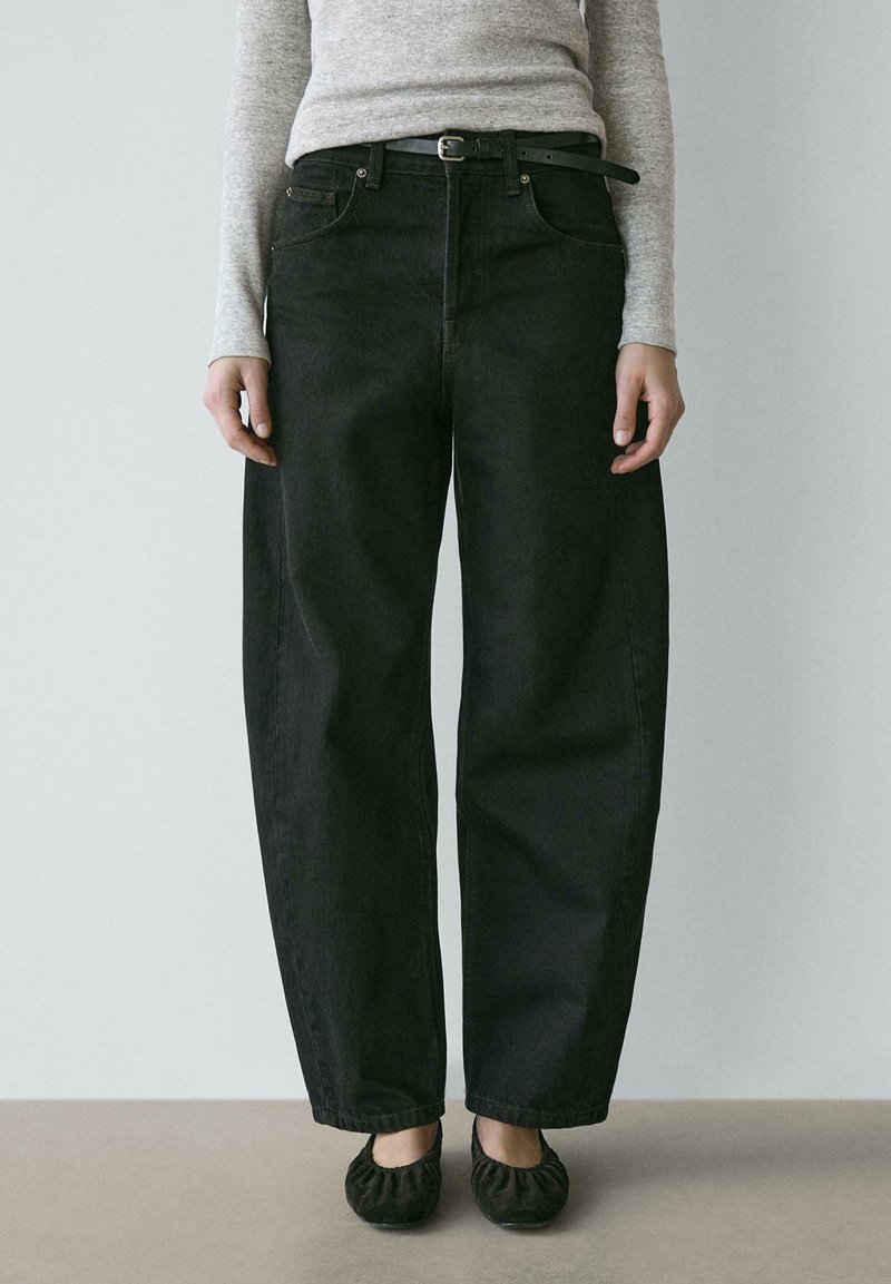 Person trägt lockere schwarze Jeans, schwarzen Gürtel, grauen langärmligen Oberteil und schwarze Ballerinas, steht vor einem schlichten hellen Hintergrund.