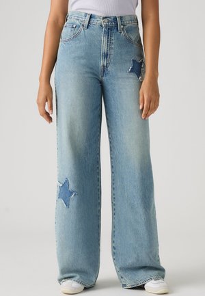 Jean large bleu clair avec des patchs en jean en forme d’étoile sur la partie supérieure de la cuisse et le bas de la jambe, porté avec des baskets blanches et un top blanc.