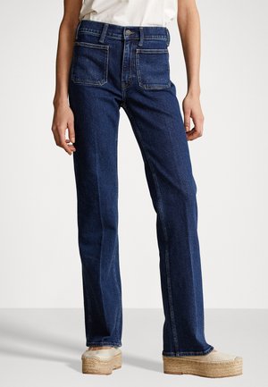 Persona che indossa jeans blu scuro a gamba dritta con tasche applicate frontali e scarpe espadrillas beige con plateau su uno sfondo neutro.