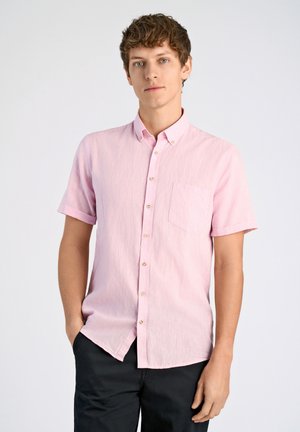 Camisa corta de manga corta en rosa claro con cuello abotonado y un único bolsillo en el pecho, hecha de una tela texturizada. Llevada con pantalones oscuros.