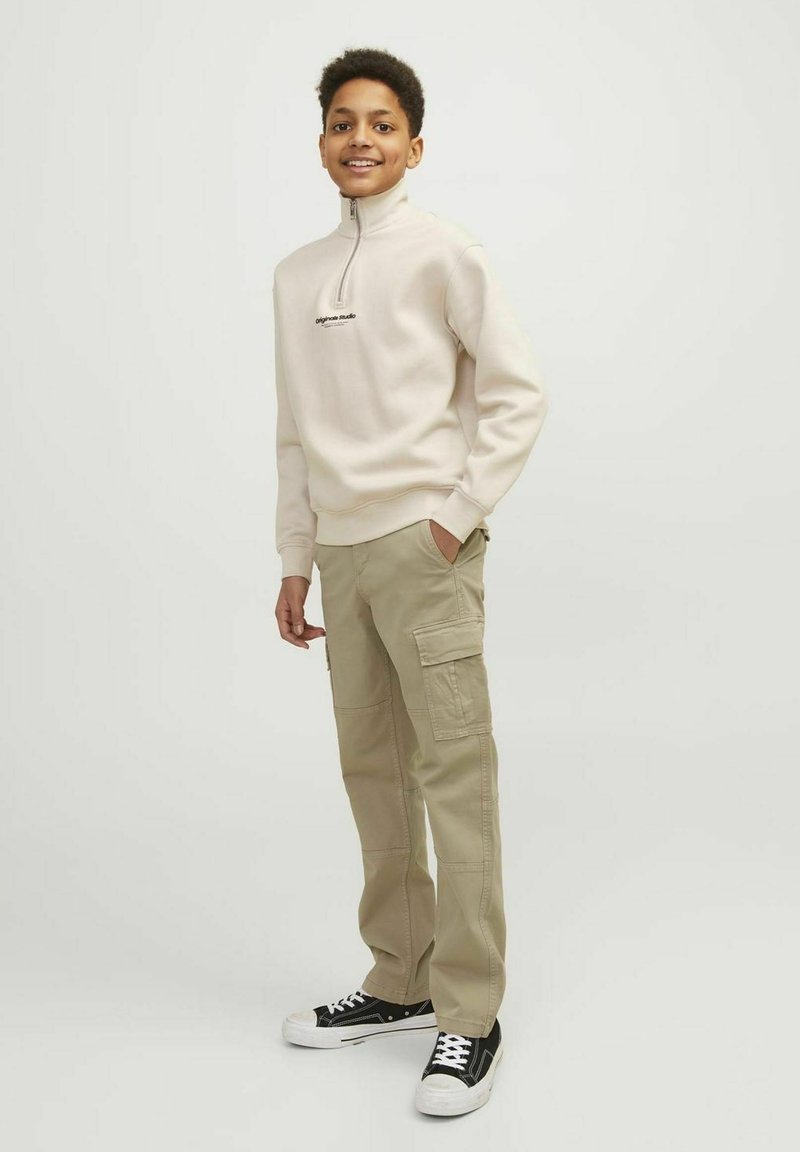 Beige fleece tröja med kvartdragkedja och svart logotyp, tillsammans med ljus khaki cargobyxor med sidofickor. Modellen har svart sneakers.