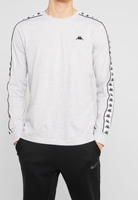 Světle šedá košile s dlouhými rukávy vyrobená z bavlny, s černými logovými akcenty na ramenou a rukávech, klasickým kulatým výstřihem.