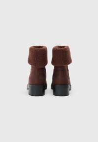 Botine din suede maro la glezna, cu o manșetă largă din blană maro texturată și o talpă robustă din cauciuc negru. Vedere din spate care evidențiază detaliul manșetei.