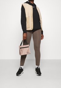 Beige ärmlös väst över svart huvtröja, bruna leggings och svarta sneakers. Rosa väska med svarts dragkedja, hålls i handen. Släta och texturerade ytor.