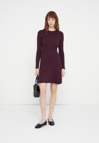 Kroppsnära, ribbad klänning i djupt burgundyröd med långa ärmar, rund halsringning och sidofickor. Matchad med en svart handväska och flats.