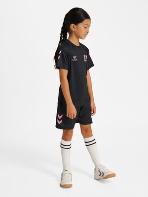 Joven niña con trenzas, vestida con un conjunto deportivo negro y rosa, calcetines blancos hasta la rodilla con rayas negras y zapatillas blancas, de pie de lado.