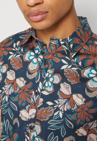 Gros plan d'une personne portant une chemise bleu foncé boutonnée avec des motifs floraux orange, blanc et bleu sarcelle et un col pointu.
