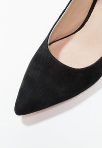 Gros plan d'une chaussure plate en daim noir à bout pointu avec une doublure intérieure beige sur fond blanc.