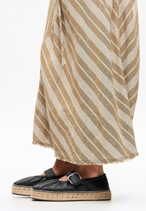 Espadrillas nere con punta tonda e dettaglio a fibbia. Indossate con una lunga gonna a righe marrone chiaro, strutturata e con orlo sfilacciato.