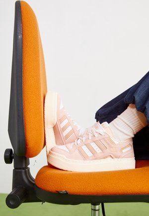 Różowe zamszowe sneakersy Adidas z białymi paskami i masywną podeszwą. Prezentowane na pomarańczowym krześle biurowym.