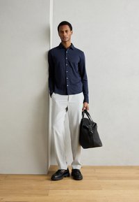 Camicia blu navy con bottoni, pantaloni grigi chiari, borsa nera con manici e scarpe nere chunky su un pavimento in legno contro una parete neutra.