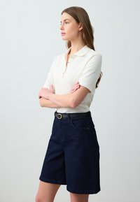 Polo a coste di colore crema con maniche corte, abbinato a pantaloni shorts in denim blu scuro e una cintura nera con fibbia dorata.