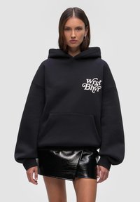 Czarna bluza z kapturem, wykonana z mieszanki bawełny, z przednią kieszenią i białym nadrukowanym logo na klatce piersiowej. W połączeniu z błyszczącą czarną mini spódnicą.