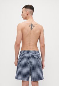 Marineblå og hvite rutete badeshorts med elastisk livbånd og baklomme, sett fra baksiden. Hud synlig på overkroppen.