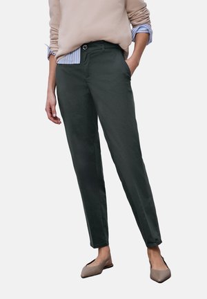 Persona che indossa pantaloni verde scuro a gamba dritta, maglietta beige a maniche lunghe con le maniche arrotolate e ballerine taupe con punta a mandorla.