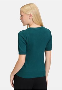 Vrouw met recht, blond haar, gekleed in een teal trui met korte mouwen en zwarte broeken, met de rug naar ons gekeerd en een zijprofiel zichtbaar.