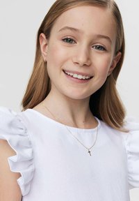 Biała bluzka z falbankowymi rękawami, noszona z delikatnym złotym naszyjnikiem z małym krzyżykiem. Tło jest minimalistyczne i neutralne.