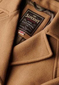 Brązowy wełniany płaszcz z wyraźnym kołnierzem, z czarną metką z napisem "SUPERDRY PREMIUM FINE TAILORING CO." oraz szczegółem w postaci brytyjskiej flagi.