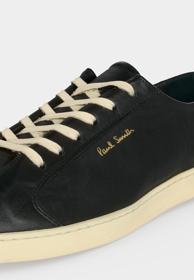 Baskets en cuir noir avec lacets et semelle couleur crème, avec le logo doré « Paul Smith » sur le côté.