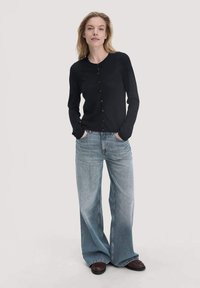 Schwarze, langärmelige Strickjacke mit Rundhalsausschnitt und Knopfleiste, kombiniert mit hellblauen, weiten Jeans und dunklen Schuhen. Weiche Stofftextur.