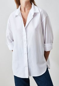Camisa blanca de botones con un corte relajado, mangas enrolladas y una textura suave y lisa. Presenta un cuello clásico y un dobladillo redondeado.