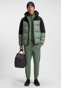 Žalia ir juoda paminkštinta striukė su gobtuvu. Derančios žalios sportinės kelnės ir juodi sportbačiai. Laiko pilką kelioninį krepšį. Paprastas dizainas.