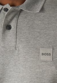 Pelēkā polo krekls, izgatavots no teksturētas auduma, ar klasisku apkakli, divām melnām pogām un kvadrātveida "BOSS" logo uzlīmi pelēkā krāsā.