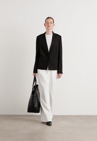 Blazer nero sopra una maglietta bianca, abbinato a pantaloni bianchi morbidi. Una borsa tote nera è in mano. Tessuti lisci, design strutturato, colori neutri.