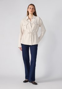 Crème-kleurig langslipshirt met twee borstzakken en een trekkoord in de taille, gecombineerd met donkerblauwe uitlopende jeans en platte schoenen.