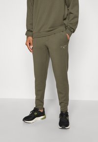 Olivgrüne Jogginghosen aus einem weichen Baumwoll-Mix, mit einem elastischen Bund, Seitentaschen und dezentem Branding in der Nähe der Hüfte.
