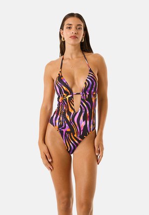 Donna che indossa un costume intero con collo all'americana con strisce zebrati nere, viola e arancioni e un taglio frontale, in piedi su sfondo bianco.