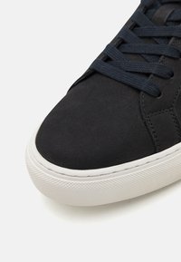 Black suede sneaker with a white rubber sole, featuring flat navy laces, a rounded toe, and visible stitching details along the edges.