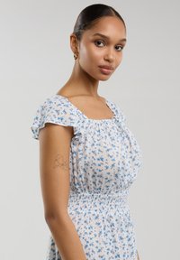 Vestido blanco con estampado floral azul, mangas cortas con volantes y cintura fruncida. Cuenta con un escote cuadrado y una tela suave y texturizada.