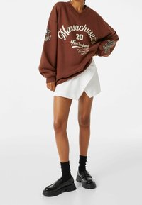 Sweat-shirt marron surdimensionné avec les inscriptions « Massachusetts » et « New England », assorti à une jupe portefeuille blanche et des mocassins à plateformes noirs portés avec des chaussettes.