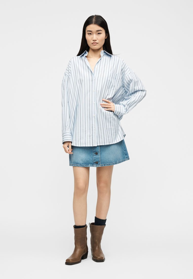 Jeune femme debout, une main sur la hanche, portant une chemise blanche oversize à rayures noires, une jupe en jean bleue, des chaussettes noires et des bottines marron.