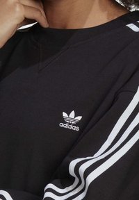 Svart sweatshirt med vita tre-randsdetaljer på ärmarna och en broderad Adidas-logotyp på vänster bröst. Mjuk tygkvalitet.