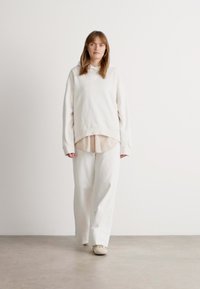 Woman walking indoors wearing an oversized white hoodie, beige shirt underneath, wide white pants, and light-colored shoes on a plain background.
