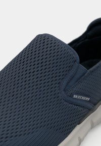 Morske plave slip-on cipele s mrežastim gornjim dijelom, teksturiranom površinom za prozračnost i fleksibilnom potplatom. Sadrže "Skechers" logo sa strane.