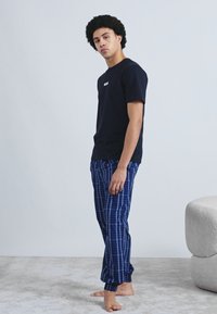 Tricou negru cu mâneci scurte și logo "BOSS", asortat cu pantaloni de pijama în carouri albastre. Modelul stă desculț pe un covor gri deschis. Fundal minimalist.