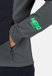 Grå atletisk topp med svarta detaljer, med en ficka med dragkedja och en grön logotyp som säger "HERO ATHLETE" på ärmen. Smetig struktur.