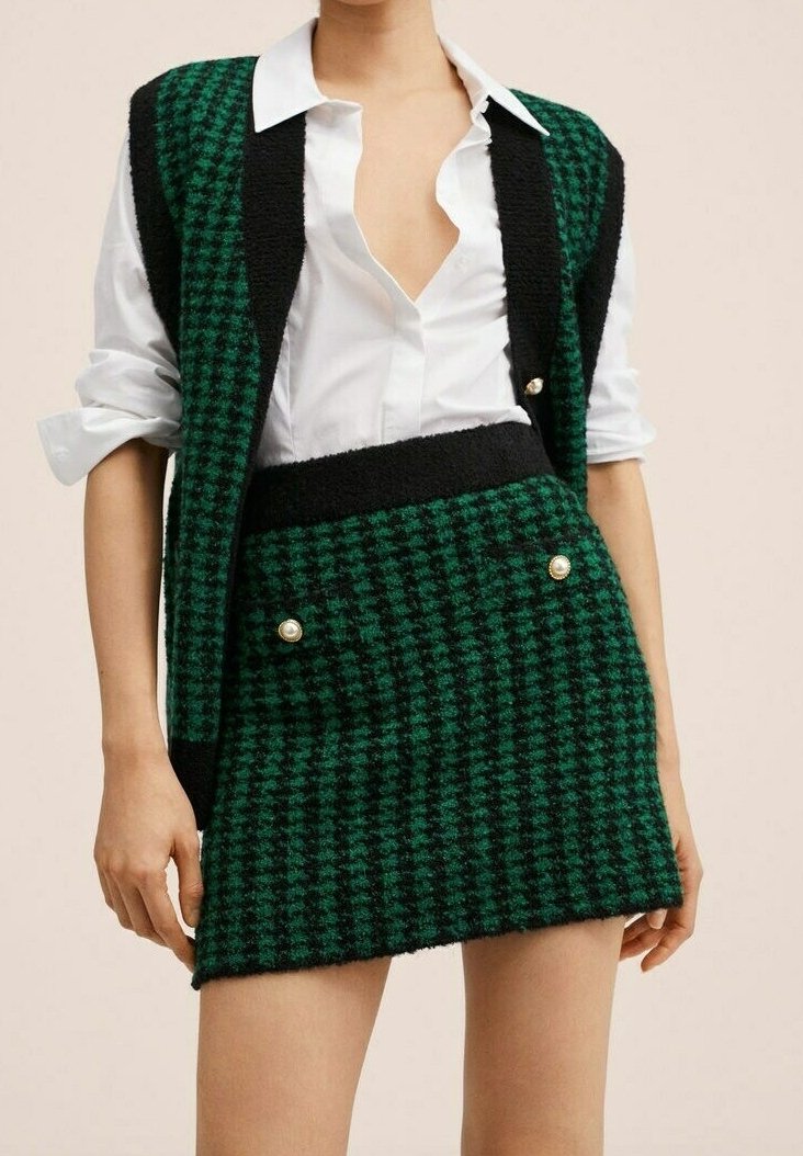 Femme portant une jupe à motif pied-de-poule vert et noir et un gilet assorti sur une chemise blanche boutonnée avec les manches retroussées.