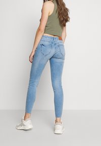 Jeans ajustados de color azul claro hechos de mezclilla, con cinco bolsillos y un parche de cuero marrón en la cintura, combinados con zapatillas blancas.