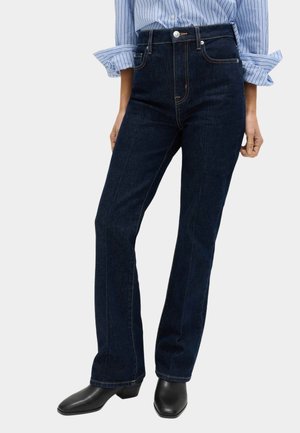 Person iført mørkeblå jeans med lige ben, sorte sko med firkantet tå og en blå stribet skjorte med opkærmede ærmer.